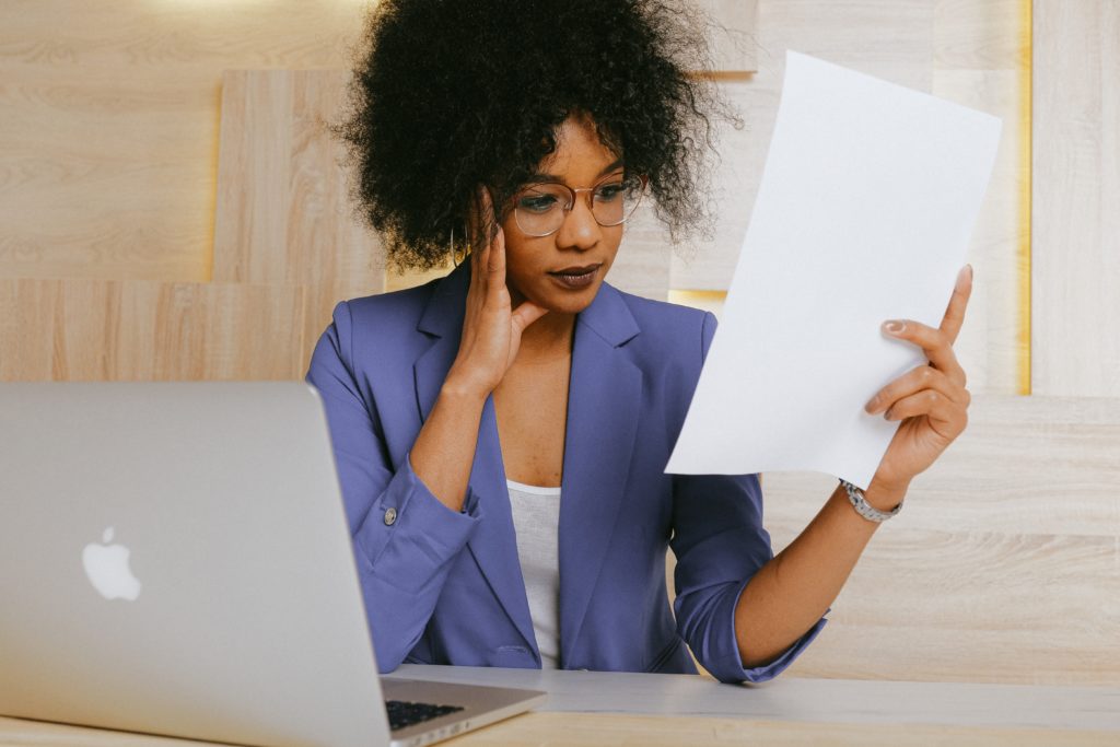 Black woman in blue blazer sitting at an apple laptop looking at a piece of paper.
