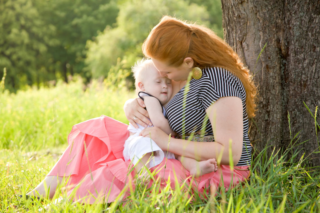 Redheaded woman holding baby with Down Syndrome in a field of grass.