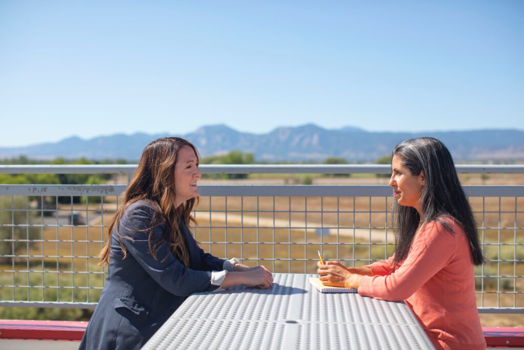 Two women in conversation at outdoor table with mountains in background.