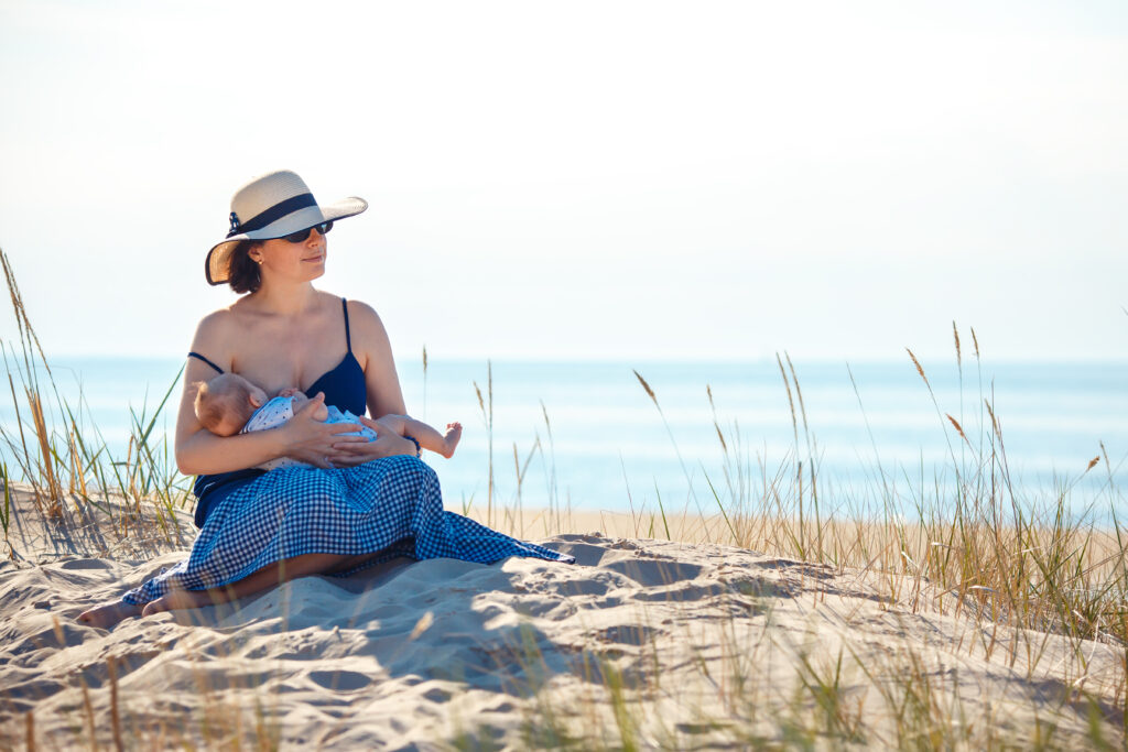 Woman in straw hat sitting on sunny beach breastfeeding.