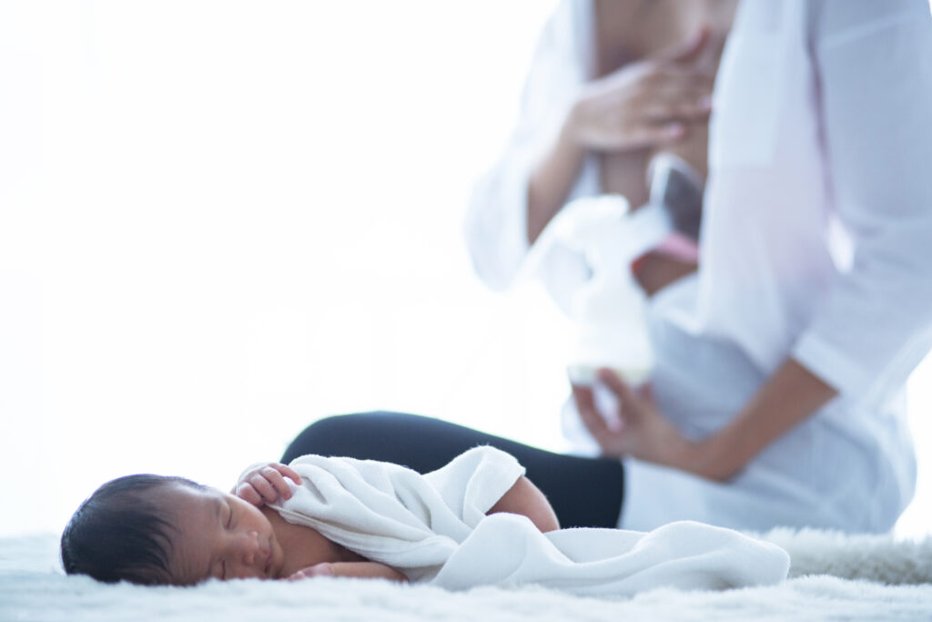 Mom on bed pumping breast milk coping with sore nipples while newborn sleeps near her.