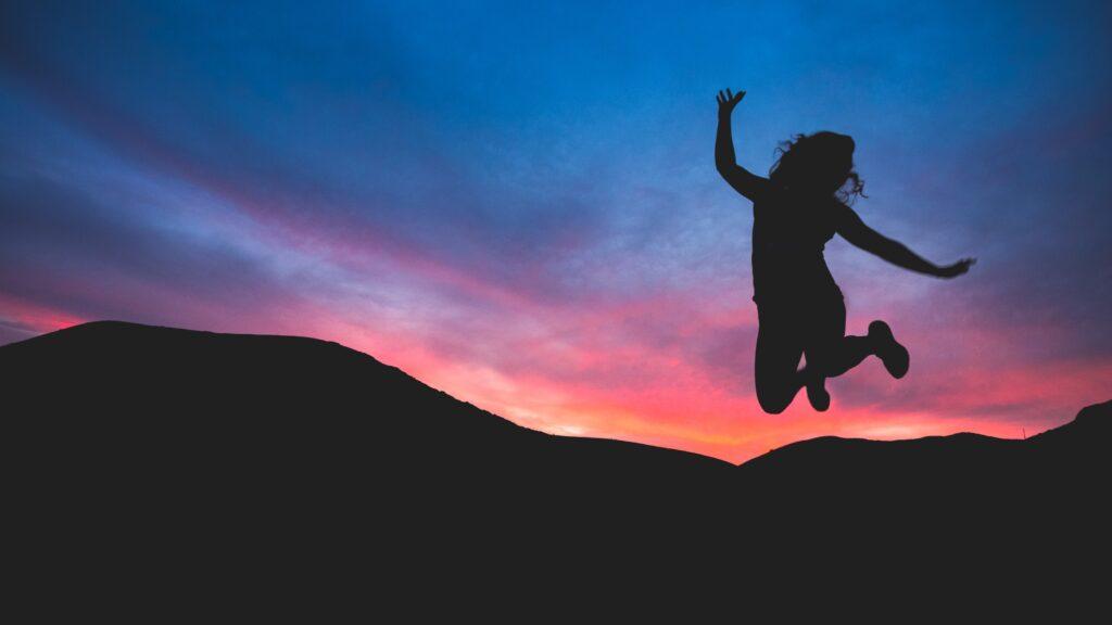 Woman in silhouette jumping over mountains with sunset in background.