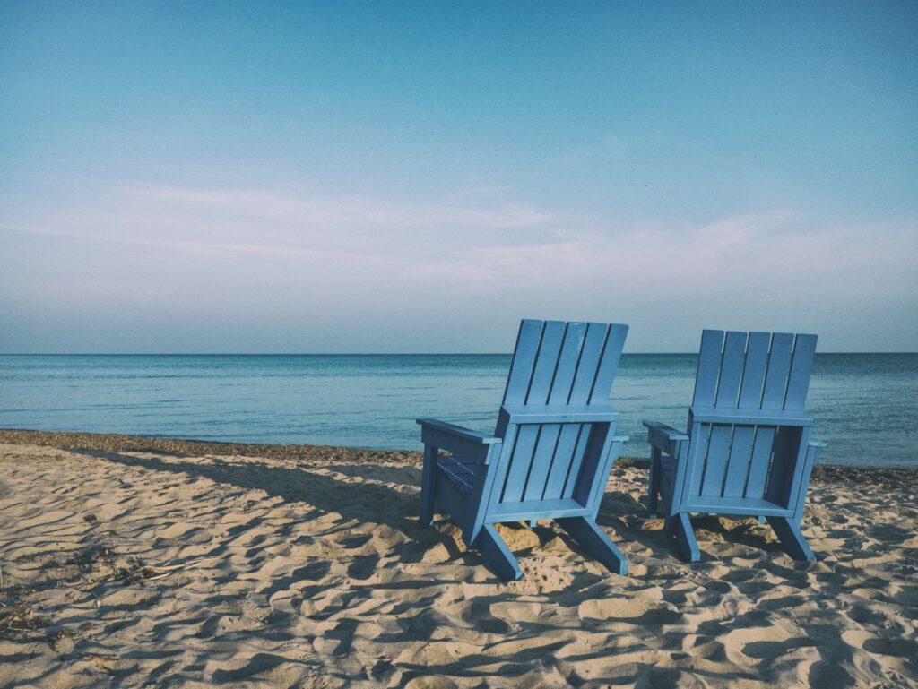 Two blue chairs sitting on sandy beach looking over body of water.