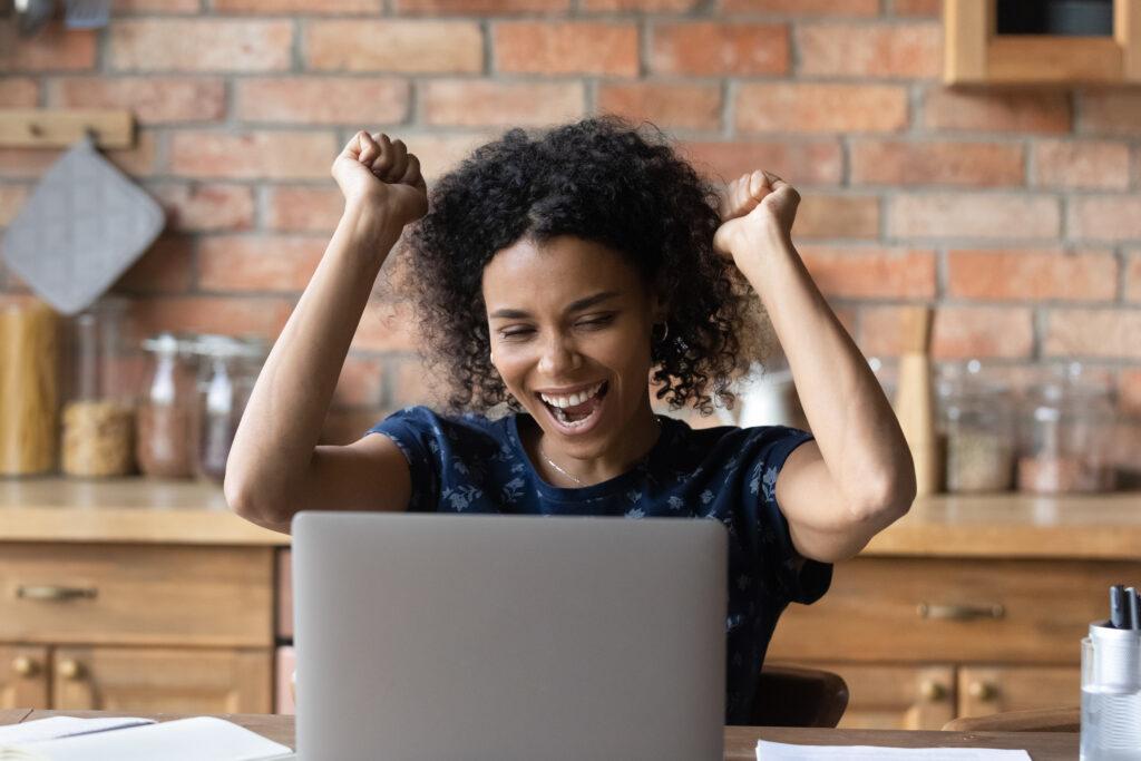 Woman raising her hands in celebration in front of a laptop computer.