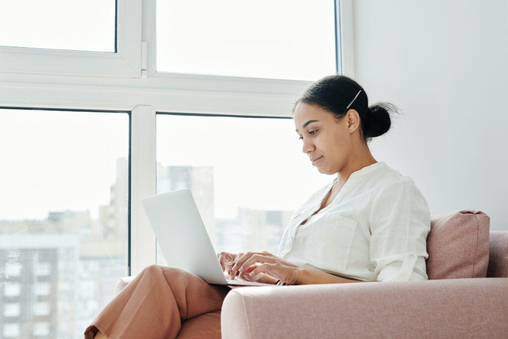Woman sitting on pink couch with laptop next to window.