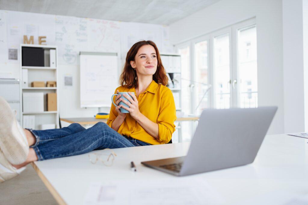Woman wearing yellow shirt holding a mug with her feet up on desk with laptop computer.