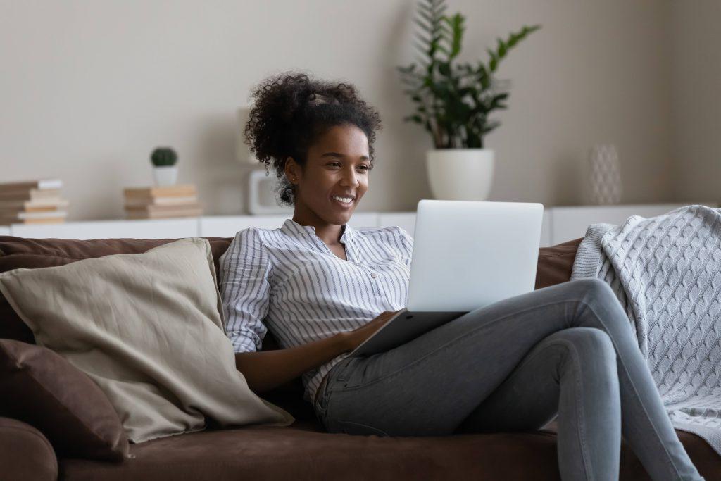 Smiling Black woman on couch balancing laptop.