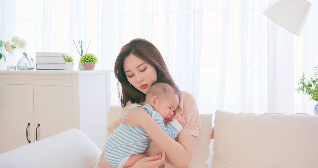 Concerned woman holds crying baby in striped onesie in bright room.
