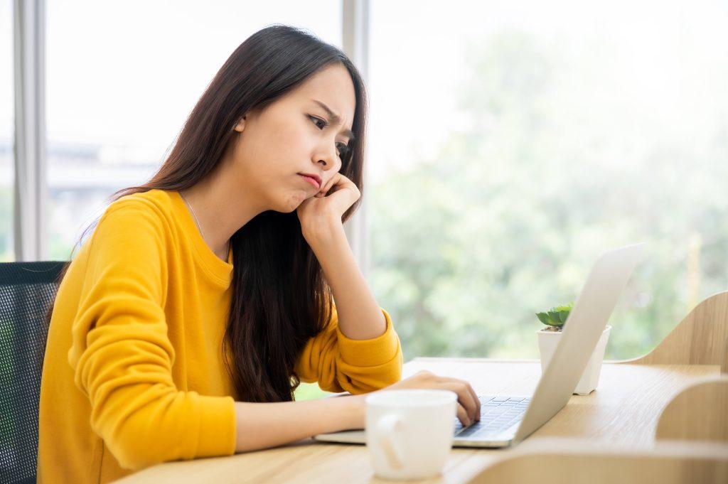 Woman in yellow sweater frowning at a computer screen with mug on desk.