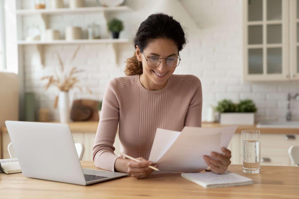 Smiling brunette with laptop computer looking at documents at a table.