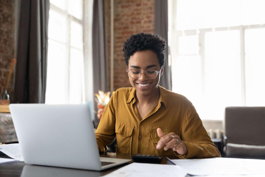 Woman in gold shirt sitting at laptop computer with calculator.