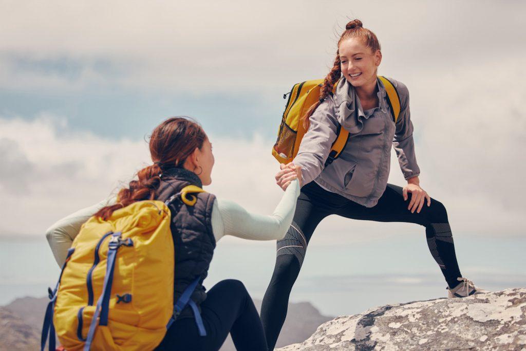 Woman getting help from a friend reach the top of the mountain while hiking.