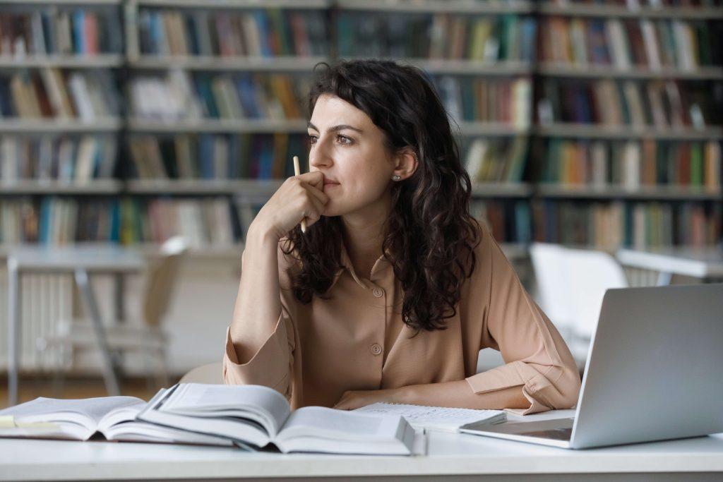Brunette woman sitting at table in libary surrounded by books and laptop computer looking into distance.