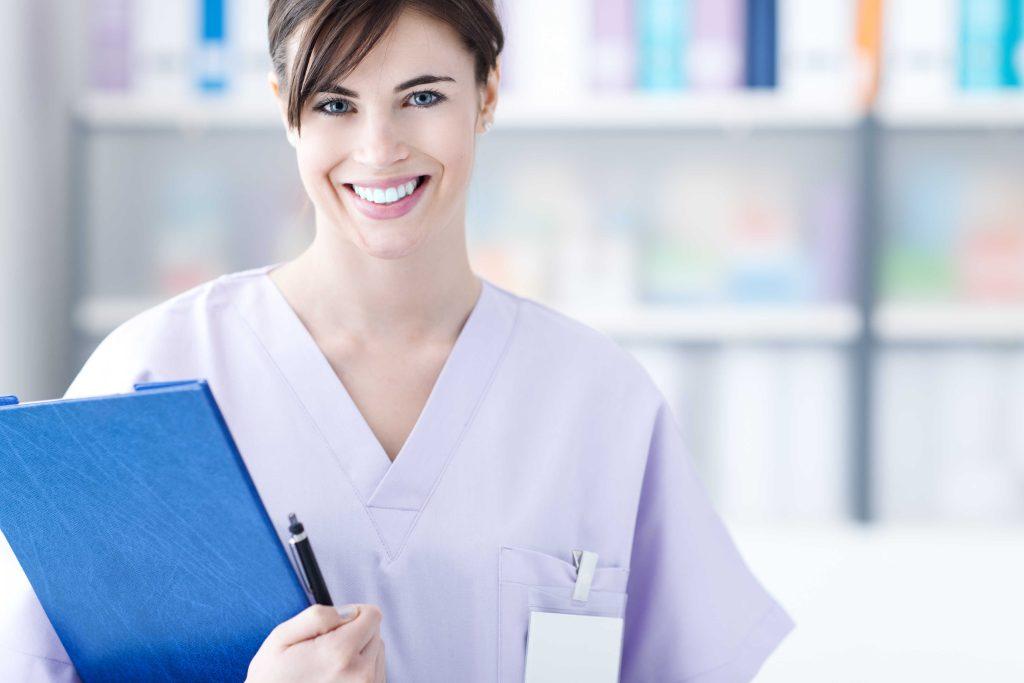 Smiling woman in lavender scrubs holding a clipboard.