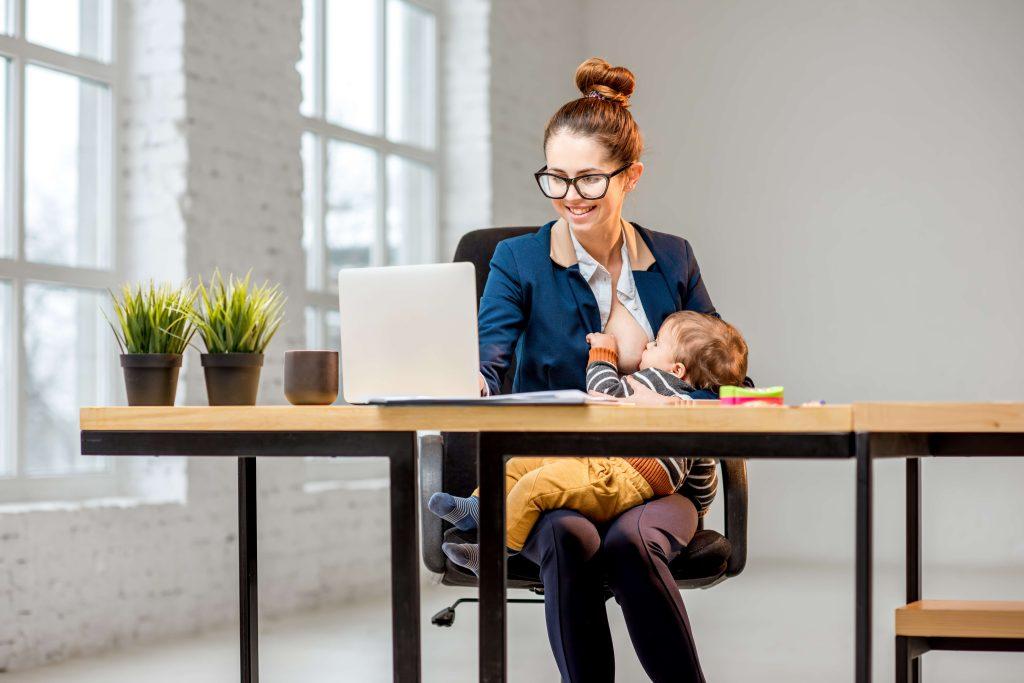 Woman at desk sits in front of laptop computer while breastfeeding.