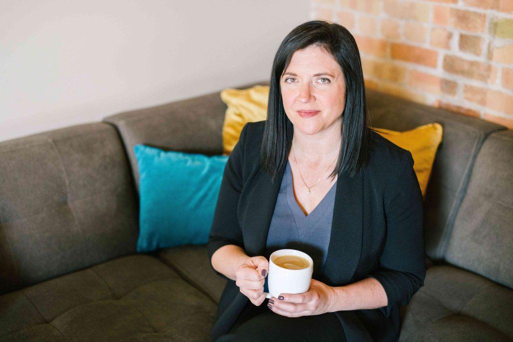 Woman sitting on couch with yellow and turquoise pillows with brick wall behind, holding a cup of coffee.