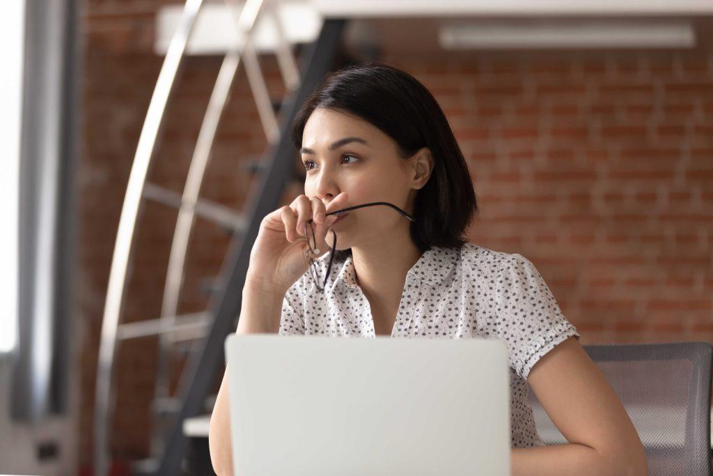 Woman looking off into distance holding eyeglasses behind a laptop computer.