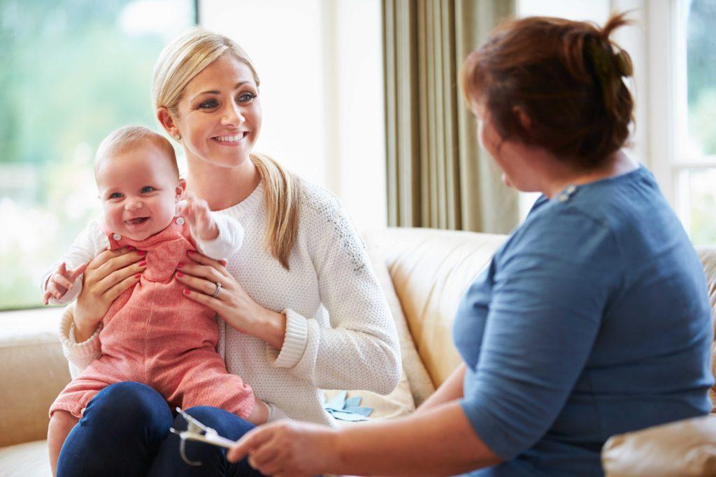 Smiling blonde woman holding baby in pink with a woman in blue sitting nearby. 